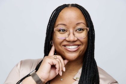 Ashli Johnson wearing glasses with her hair in braids with center part. She has on a blush colored blouse, watch, necklace, and earrings.